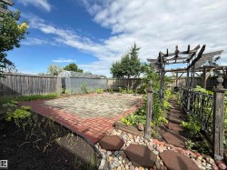 Expansive backyard featuring a red brick paver patio, wood privacy fencing, a transparent greenhouse structure, and a rustic wooden pergola with decorative metal railings - 