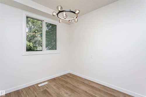 Room featuring a contemporary circular chandelier, a window with exterior tree scenery, wood-finish flooring, white walls, and white baseboards - 5418 144B Avenue, Edmonton, AB 