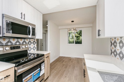 Modern kitchen featuring stainless steel appliances, geometric pattern tile backsplash, light wood-finish flooring, white cabinetry with black hardware, and a separate dining area with a contemporary chandelier - 5418 144B Avenue, Edmonton, AB 