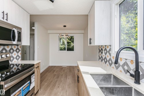 Modern kitchen featuring white cabinetry, light countertops, stainless steel appliances, a geometric tile backsplash, and wood-finish flooring - 5418 144B Avenue, Edmonton, AB 