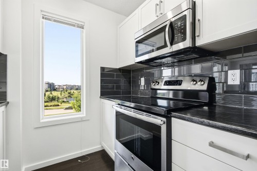 Modern kitchen featuring white cabinetry, dark countertop, stainless steel appliances, dark subway tile backsplash, and a tall window - 435 5515 7 Avenue, Edmonton, AB 