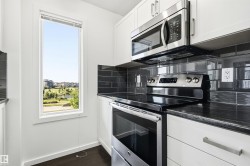 Modern kitchen featuring white cabinetry, dark countertop, stainless steel appliances, dark subway tile backsplash, and a tall window - 
