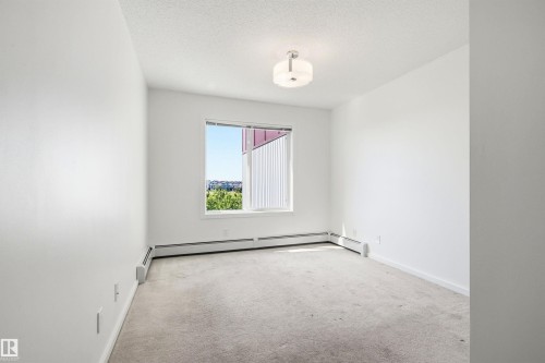 Neutral-toned carpeted room featuring a window with exterior residential and green landscape, white painted walls, white baseboards, and a semi-flush ceiling light fixture - 435 5515 7 Avenue, Edmonton, AB 