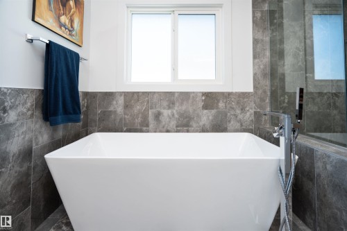 Freestanding soaking tub with a floor-mounted chrome faucet, surrounded by gray stone-patterned wall tiles - 4714 Charles Bay Bay, Edmonton, AB 