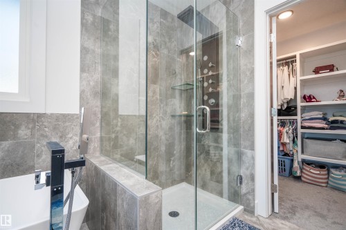 Contemporary bathroom featuring a frameless glass shower enclosure with rain shower head, full-height stone-finish wall tiling, separate soaking tub with a modern floor-mounted fixture, and a built-in walk-in closet with integrated shelving - 4714 Charles Bay Bay, Edmonton, AB 