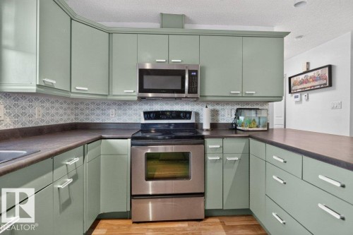 Kitchen featuring sage-toned cabinetry, stainless steel appliances, patterned tile backsplash, and wood-finish flooring - 21320 90 Avenue, Edmonton, AB 