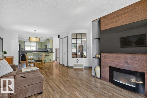 Open-concept living space featuring wood-finish flooring, a contemporary fireplace with a wood-finish surround, and a kitchen with light green cabinetry - 21320 90 Avenue, Edmonton, AB 