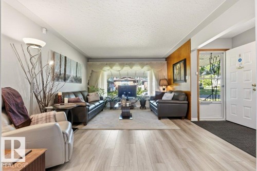 Spacious living area featuring light wood-finish flooring, a large front-facing window, and a white paneled entry door with a glass insert - 10235 74 Street, Edmonton, AB 