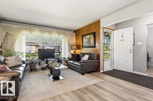 Living area featuring wood-finish flooring, a large picture window, and a paneled entry door with a storm door - 10235 74 Street, Edmonton, AB 