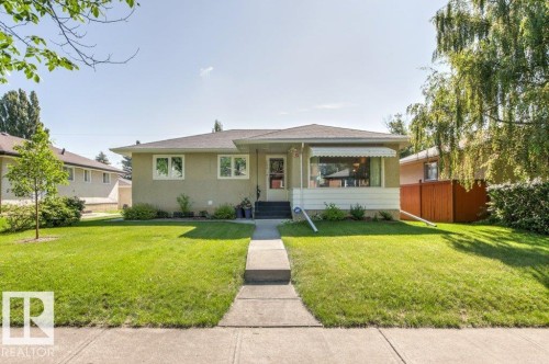 Ranch-style residence featuring a stucco exterior, a large front lawn, a concrete walkway, established landscaping, and a bay window with an awning - 10235 74 Street, Edmonton, AB 