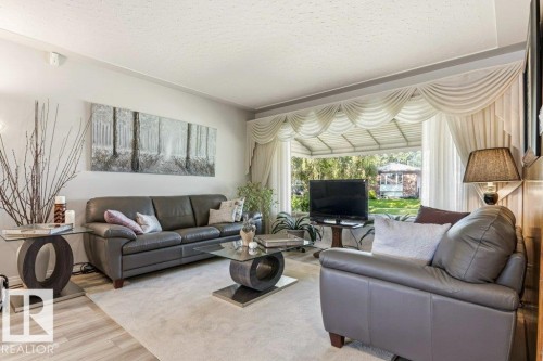 Spacious living area featuring light-toned wood-finish flooring, a plush area rug, and a large bay window - 10235 74 Street, Edmonton, AB 