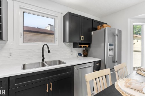 Modern kitchen featuring black cabinetry, light countertops, a stainless steel double-basin sink with a matte black faucet, a white subway tile backsplash, and stainless steel appliances - 15252 81 Street, Edmonton, AB 