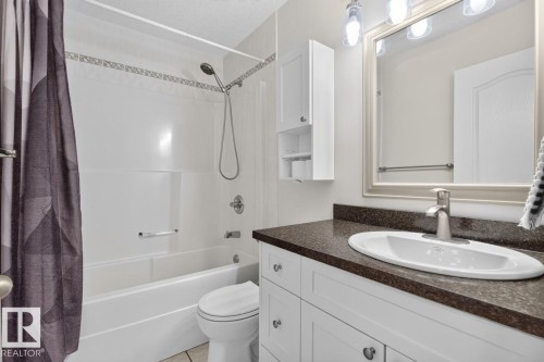 Full bathroom featuring a white bathtub with a showerhead, a toilet, a vanity with a dark countertop and an undermount sink, and a framed mirror with overhead lighting - 619 Layton Crest, Edmonton, AB 