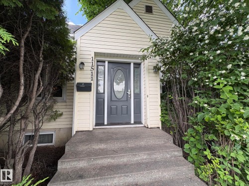 Cream-colored siding facade featuring a gray front door with an oval-shaped glass insert, concrete steps, and integrated address numbers - 11511 101 Street, Edmonton, AB 