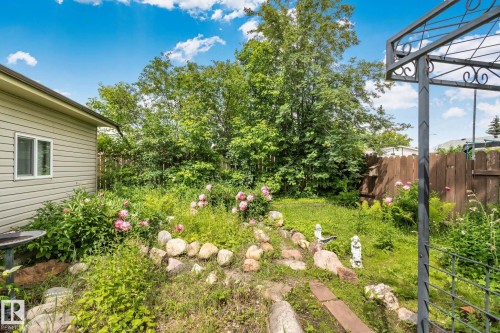 Lush green backyard featuring established trees, flowering shrubs, a rock-lined garden bed, a wooden privacy fence, and an ornamental metal arbor - 30 Garden Valley Drive, Stony Plain, AB - Outdoor