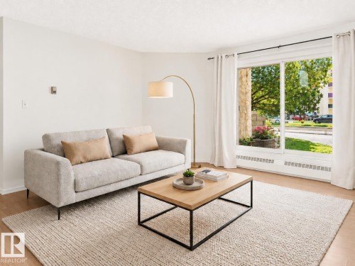 Bright living space featuring a large picture window, light wood-finish flooring, and white painted walls - 143 10636 120 Street, Edmonton, AB 