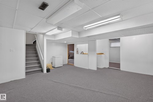 Spacious lower-level room featuring gray carpet flooring, white paneled walls, and a dropped ceiling with fluorescent lighting fixtures - 3841 62 Street, Edmonton, AB 
