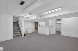 Spacious lower-level room featuring gray carpet flooring, white paneled walls, and a dropped ceiling with fluorescent lighting fixtures - 