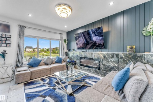 Spacious living area featuring large windows with a water backdrop, modern fireplace, marble-finish wall accents, recessed lighting, and a contemporary ceiling fixture - 5809 65 Street, Beaumont, AB - Indoor Photo Showing Living Room With Fireplace