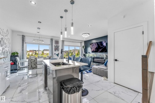 Open-concept living area featuring large format white tile flooring, a kitchen island with an undermount sink and contemporary pendant lighting, a dining area with expansive windows, and a living room with a feature wall and fireplace - 5809 65 Street, Beaumont, AB - Indoor Photo Showing Kitchen With Fireplace