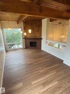 Spacious living area featuring wood-finish flooring, exposed wood beams, and a prominent brick fireplace with a shiplap-style surround - 