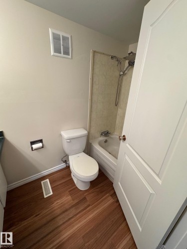 Bathroom featuring wood-finish flooring, a white toilet, a bathtub with a showerhead, a wall-mounted ventilation unit, and a paneled white door - 482 Lee Ridge Road Nw, Edmonton, AB 