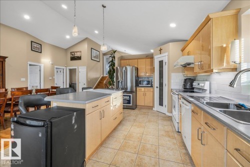Open-concept kitchen featuring light wood-finish cabinetry, tile flooring, a central island with countertop seating, stainless steel appliances, and vaulted ceilings with recessed lighting - 6050 164A Avenue, Edmonton, AB - Indoor Photo Showing Kitchen