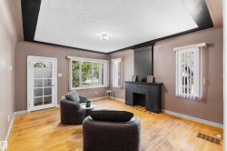 Living area featuring wood-finish flooring, a decorative fireplace with a dark mantelpiece, multiple windows with vertical blinds, a glass-paneled entry door, and a tray ceiling with recessed lighting - 