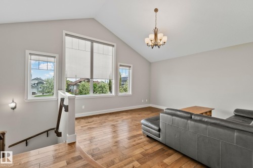 Vaulted ceiling living area featuring wood-finish flooring, a contemporary chandelier, large windows with roller shades, and a prominent staircase with a dark wood handrail - 9 Greenfield Wynd, Fort Saskatchewan, AB - Indoor Photo Showing Other Room
