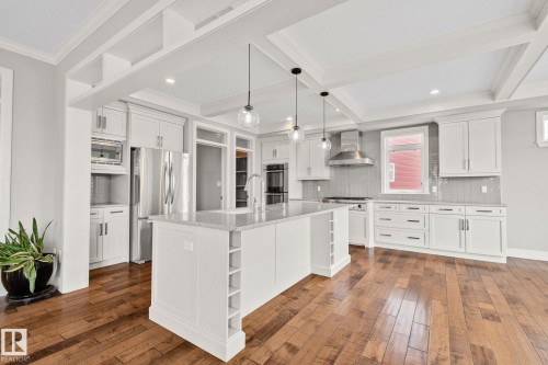 Spacious kitchen featuring an expansive center island with integrated wine storage, white cabinetry, stainless steel appliances, wood-finish flooring, and a coffered ceiling - 9 Greenfield Wynd, Fort Saskatchewan, AB - Indoor Photo Showing Kitchen With Stainless Steel Kitchen With Upgraded Kitchen