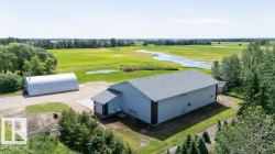 Expansive property featuring a large light-colored commercial-style building with a dark-trimmed base, an additional quonset hut structure, and surrounding green landscapes with mature trees - 