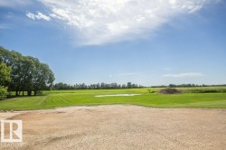 Expansive natural landscape featuring a gravel foreground, a broad green lawn, a cluster of mature trees, a small body of water, and distant foliage - 