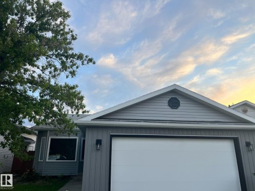 Gray siding exterior featuring a white garage door, large picture window, black exterior lighting fixtures, and a decorative gable vent - 3320 43 Street, Edmonton, AB 