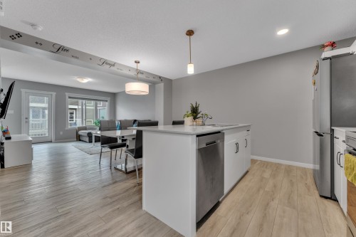 Open-concept living space featuring wood-finish flooring, a kitchen island with integrated sink, stainless steel dishwasher, and a contemporary pendant light fixture - 82 1816 Rutherford Road, Edmonton, AB 
