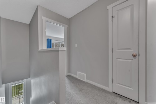 Carpeted hallway featuring light gray walls, white trim, a solid white door with a round brass-tone knob, and an architectural half-wall with an interior window - 82 1816 Rutherford Road, Edmonton, AB 
