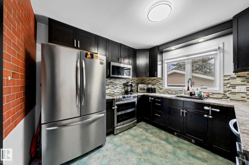 Kitchen featuring dark wood-finish cabinetry, stainless steel appliances, a mosaic tile backsplash, and a double basin sink - 5418 17A Avenue, Edmonton, AB 