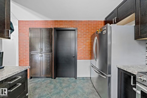 Kitchen area featuring a brick accent wall, a stainless steel refrigerator, dark wood-finish cabinetry, light-colored countertops, and gray tile flooring - 5418 17A Avenue, Edmonton, AB 