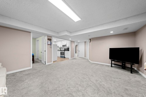 Expansive carpeted area with recessed lighting, complemented by a kitchen space featuring white cabinetry, a built-in oven, and a refrigerator - 5418 17A Avenue, Edmonton, AB 