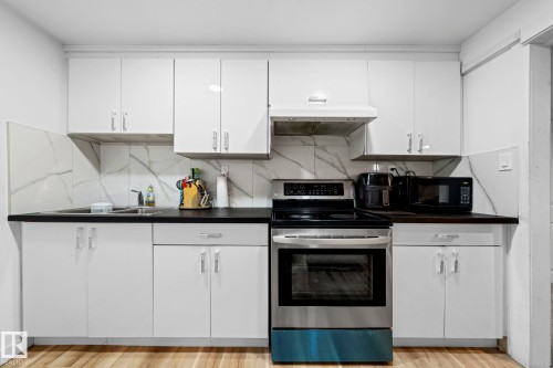 Kitchen featuring white cabinetry, dark countertops, a stainless steel electric range with an overhead vent, a stainless steel sink, and a white marble-patterned backsplash - 5418 17A Avenue, Edmonton, AB 