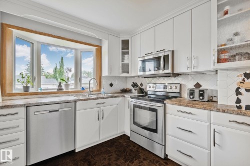 Kitchen featuring a bay window, white cabinetry, integrated stainless steel appliances, a double basin sink, and wood-finish flooring - 10715 33 Avenue, Edmonton, AB 