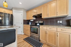 Well-appointed kitchen featuring light wood cabinetry, stainless steel appliances, granite-style countertops, dark tile backsplash, and light wood-finish flooring - 