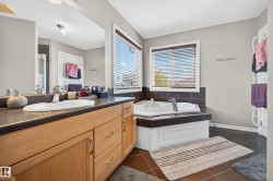 Bathroom featuring a wood-finish vanity with a built-in sink, a large wall-mounted mirror, a tiled corner bathtub, dual windows with blinds, and dark tile flooring - 