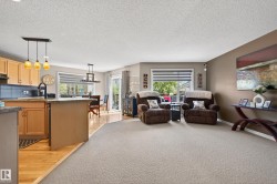 Open-concept living space featuring light wood cabinetry, a kitchen island with seating, a dining area with pendant lighting, and a carpeted living room - 