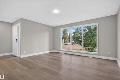 Spacious room featuring a large three-panel window, light wood-finish flooring, white baseboards, and a contemporary flush-mount ceiling light - 33 Brookside Avenue, Spruce Grove, AB 