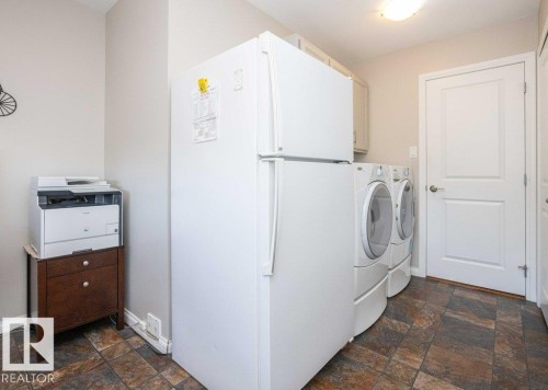 Functional utility area featuring a full-size white refrigerator, stackable white washer and dryer units, and an overhead cabinet for storage - 10420 111 Avenue, Westlock, AB 