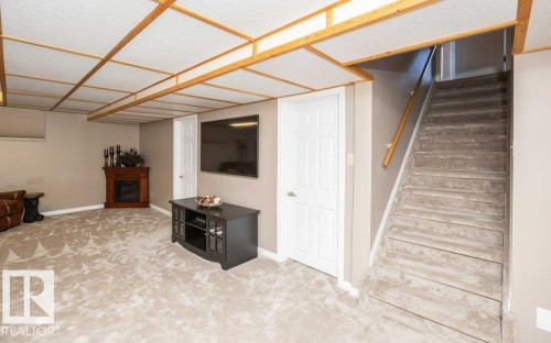 Carpeted recreation room featuring a corner fireplace with wood mantel, drop ceiling with wood framing, and a carpeted staircase with wood handrail - 10420 111 Avenue, Westlock, AB 