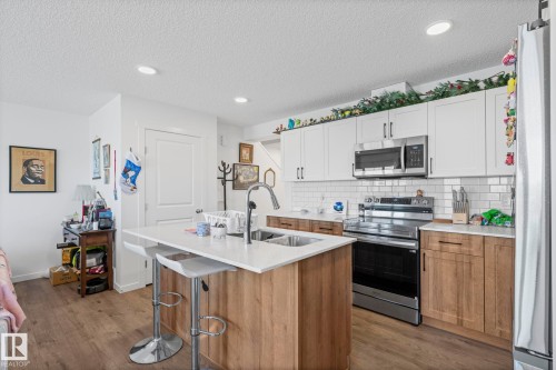 Open-concept kitchen featuring an island with seating, stainless steel appliances, two-tone cabinetry, subway tile backsplash, and wood-finish flooring - 16 19904 31 Avenue, Edmonton, AB 