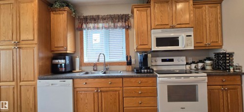 Kitchen featuring wood cabinetry, a window with blinds, a white electric range with an overhead microwave, and a stainless steel sink with a gooseneck faucet - 4221 46 Street, Vegreville, AB 