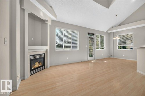 Open concept living area featuring wood-finish flooring, a contemporary fireplace with mantel, built-in shelving, vaulted ceilings, and extensive window coverage including a glass-paneled door - 20 4821 Terwillegar Common, Edmonton, AB 