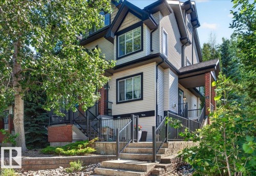 Multi-story residence featuring light-colored siding, black trim windows, a covered front porch with brick columns, and a terraced front yard with stone steps and retaining walls - 20 4821 Terwillegar Common, Edmonton, AB 